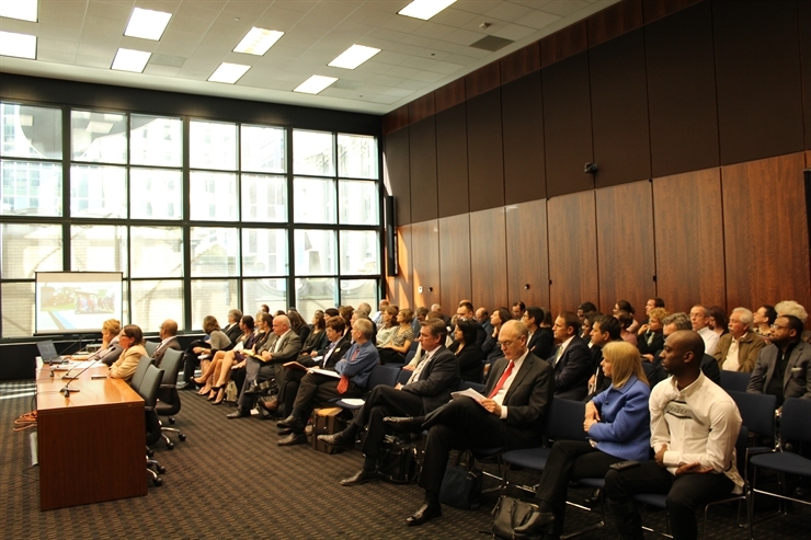 Two individuals testifying at a hearing in a crowded room